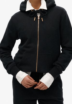 Personne portant un sweat à capuche noir zippé avec une fermeture éclair dorée et des cordons sur une chemise blanche à col, mains ajustant la fermeture éclair.