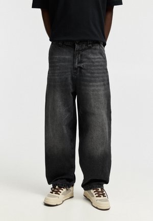 Personne portant un jean noir délavé ample et des baskets crème avec des lacets marron, debout devant un fond uni.