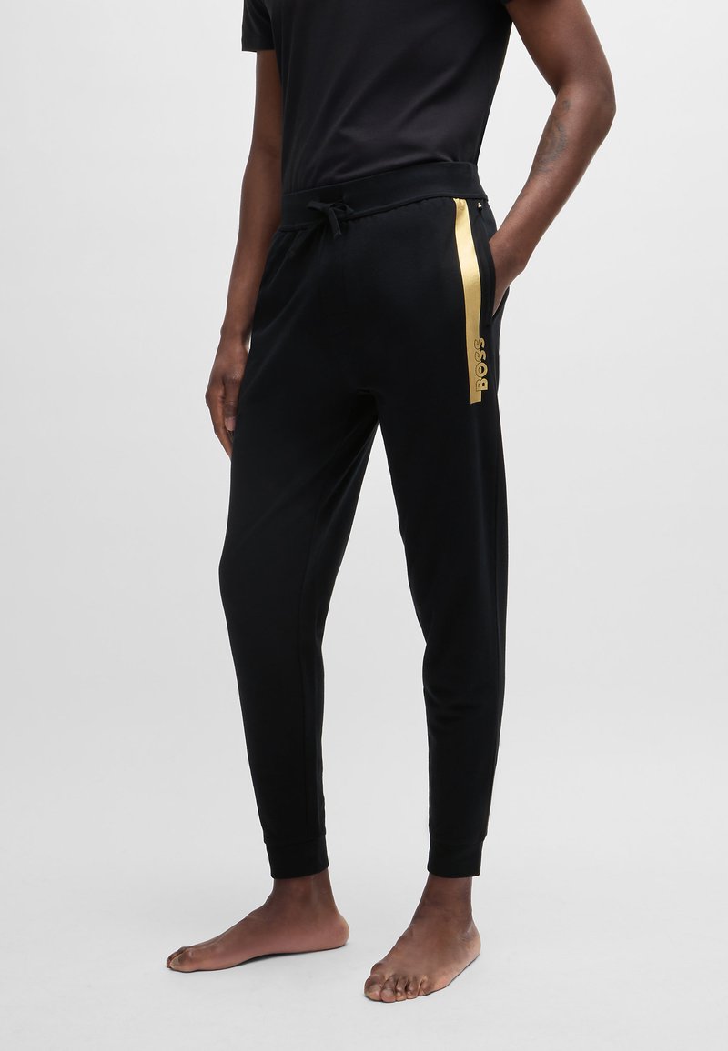 Pantalones de chándal negros de tela suave, con un ajuste slim, cinturilla elástica, cordón y una banda con logo dorado contrastante en el lado.
