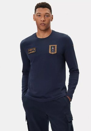 Camicia blu navy a maniche lunghe realizzata in un tessuto liscio. Presenta toppe sul lato sinistro del petto e sulla spalla, con dettagli in oro e rosso.