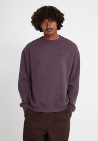 Homme aux cheveux bouclés portant un sweat-shirt violet avec un petit logo noir brodé sur le côté gauche de la poitrine, debout les mains dans les poches d'un pantalon marron.