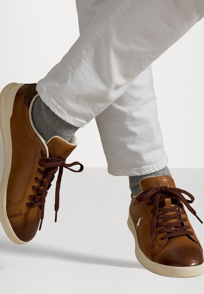 Rjave usnjene tenisice z okroglim nosom, rjavimi vezalkami in bež podplatom. Sive nogavice so vidne nad belimi hlačami. Podrobnost logotipa na strani.