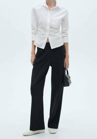 Chemise blanche boutonnée avec manches longues et une seule poche poitrine, associée à un pantalon noir taille haute à jambes larges et des chaussures de couleur crème.