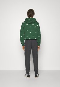 Grüne Kapuzenjacke mit weißen Logo-Mustern, kombiniert mit dunkelgrauen Jogginghosen. Die Kapuzenjacke verfügt über eine Kängurutasche und eine verstellbare Kapuze.