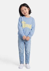 Jeune fille souriante, portant un sweat-shirt bleu clair avec un texte jaune, un jean bleu clair, et des chaussures blanches sans lacets, debout les mains jointes.