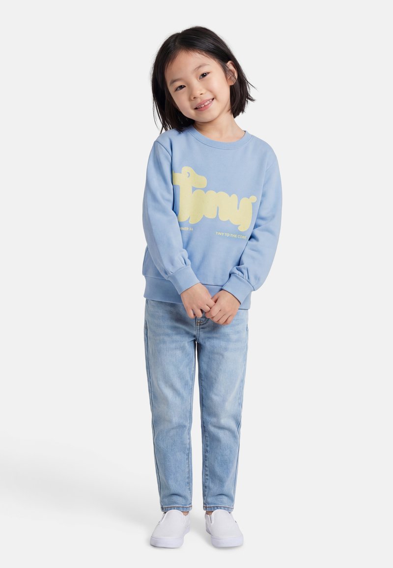 Jeune fille souriante, portant un sweat-shirt bleu clair avec un texte jaune, un jean bleu clair, et des chaussures blanches sans lacets, debout les mains jointes.