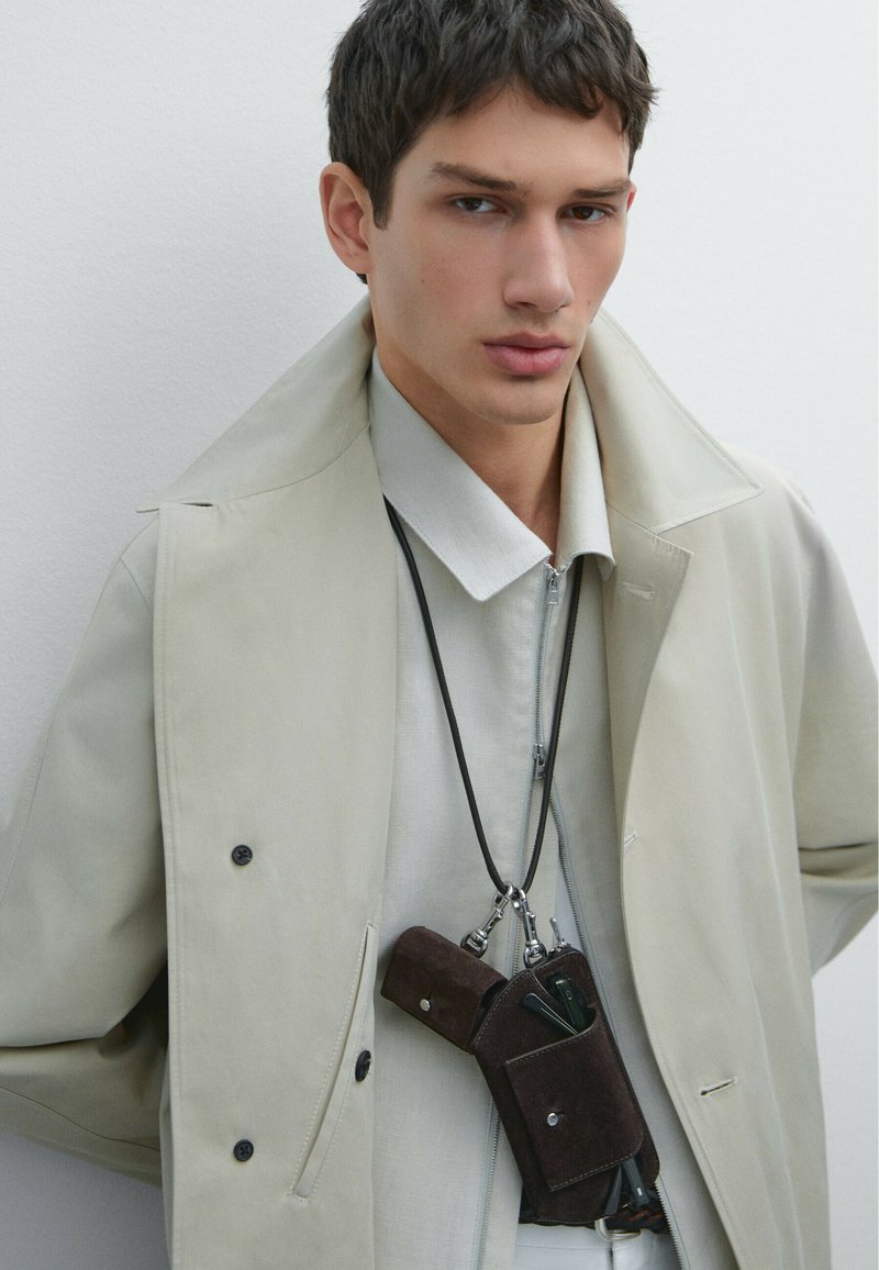 Beige trenchcoat over en lys grå skjorte, med en brun ruskinds telefonholder med en sort snor, fastgjort med metalclips.