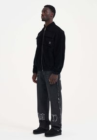 Mustat corduroy-tyyppiset vetoketjulla varustetut takki, joissa on kaksi etutaskua, yhdistettynä tummiin farkkuihin, joissa on graafinen printti ja rispaantuneet reunat.