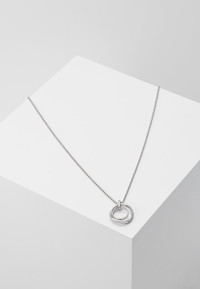 Collier en argent avec deux pendentifs circulaires irréguliers entrelacés sur une chaîne délicate, présenté sur une surface géométrique blanche.
