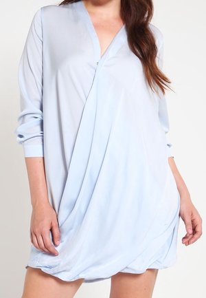 Femme portant une robe portefeuille bleu clair à manches longues, col en V et ourlet asymétrique drapé, debout devant un fond uni.