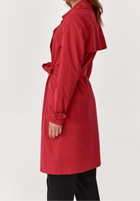 Mujer vestida con un abrigo de trench rojo largo y con cinturón, que tiene un yugo trasero y detalles de hebilla en las mangas, de pie frente a un fondo liso.