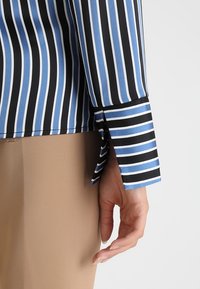 Blusa a righe con strisce orizzontali nere, blu e bianche. Include un polsino con una striscia blu a contrasto e un orlo inclinato.