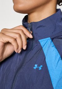 Mornarska jakna s svijetloplavim akcentima, koja ima zatvarač na ovratniku i strukturalnu tkaninu. Logo Under Armour-a je vidljiv na prednjoj strani.