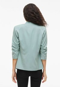 Femme aux longs cheveux bouclés portant un blazer à manches froncées vert clair et un pantalon noir, vue de dos sur fond blanc.