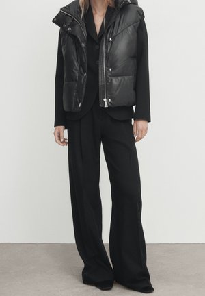 Vrouw die zwarte broek met wijde pijpen, een zwart colbert en een zwarte mouwloze gewatteerde bodywarmer met zilveren rits en knopen draagt, staand op een grijze vloer.