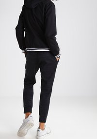 Chaqueta con capucha negra y puños de rayas blancas, combinada con pantalones jogger gris oscuro y zapatillas blancas, vista desde atrás. Tejido suave y ajuste casual.