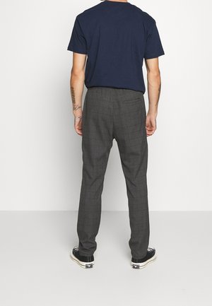 Mann, der dunkelgraue karierte Hose, ein marineblaues T-Shirt und schwarze Sneakers trägt, von hinten vor einfachem Hintergrund gesehen.