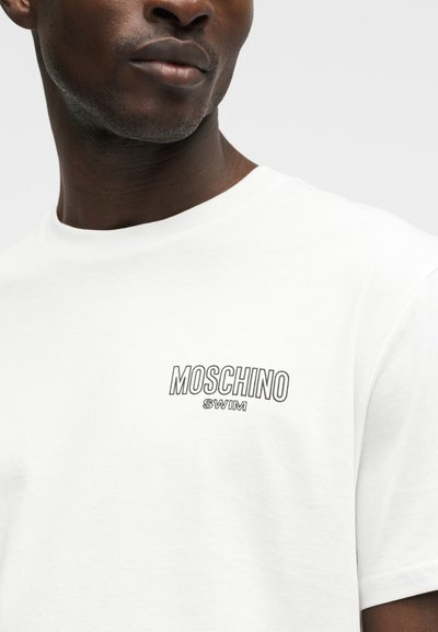 Άνδρας που φοράει λευκό μπλουζάκι με τη λογότυπο "MOSCHINO SWIM" τυπωμένο με μαύρο στο στήθος, σε απλό φόντο.