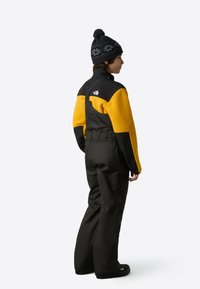 Skidställ med svarta och gula fleecemattärmar, med en svart överdel och vattentäta mörka byxor. Komplett med en svart stickad mössa med en tofs.