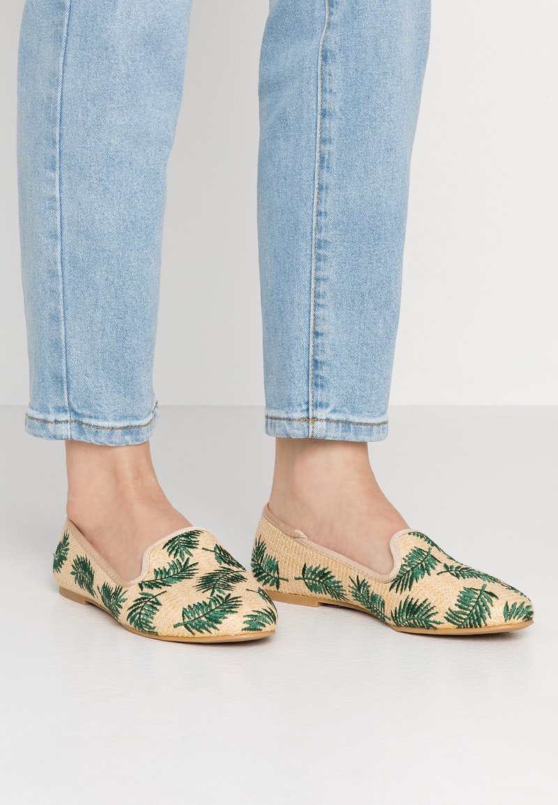 Pieds portant un jean bleu clair et des chaussures slip-on beiges avec des motifs de feuilles vertes, debout sur un sol blanc.