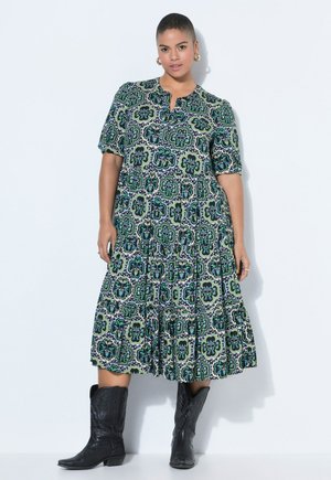 Frau trägt ein grün-schwarz-weiß gemustertes Midikleid mit kurzen Ärmeln und schwarze Cowboy-Stiefel bis zur Mitte der Wade, steht vor einer weißen Wand.