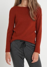 Sweter w kolorze rdzy z długim rękawem, o dekolcie w łódkę i prążkowanej struktury, zestawiony z szarymi spodniami na sznurek. Prosty i casualowy design.