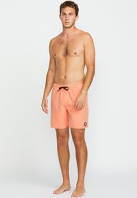 Pfirsichfarbene Badeshorts mit einem elastischen Bund und schwarzem Kordelzug, ausgestattet mit einem kleinen Logopatch am unteren linken Bein.
