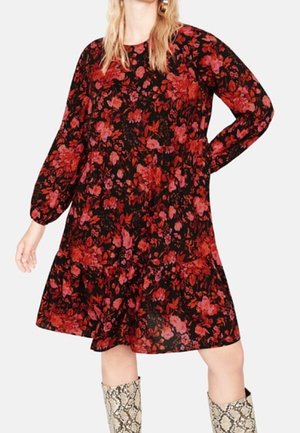 Kobieta nosząca długą sukienkę z długim rękawem do kolan w czarnym kolorze z czerwonym i różowym kwiatowym wzorem, zestawioną z wysokimi kozaczkami o wzorze skóry węża.