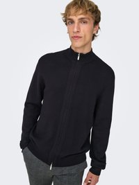 Maglione nero con zip e collo alto, realizzato in tessuto a maglia liscia e polsini a coste. Il modello indossa pantaloni a quadri per creare contrasto.