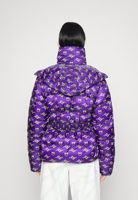 Persoon die een paarse doorgestikte jas draagt met herhaalde gele logo's, een hoge kraag en capuchon, gezien van achteren tegen een effen achtergrond.