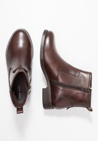 Une paire de bottes en cuir marron foncé à la cheville avec des fermetures éclair sur le côté et des talons empilés bas, présentées en vue de dessus et de côté sur un fond blanc.