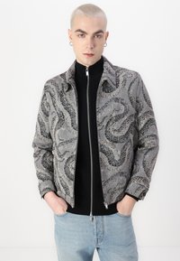 Grå zip-up jakke med et tekstureret slangeprint-design i mørkere nuancer. Den har krave og forlommer, og den er parret med en sort turtleneck.