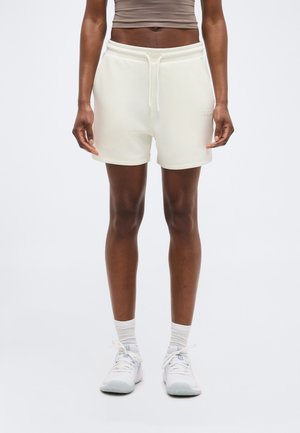 Άτομο που φοράει μπεζ σορτς με κορδόνι, λευκές κάλτσες και λευκά αθλητικά παπούτσια, στέκεται με τα χέρια χαλαρά δίπλα στο σώμα σε απλό φόντο.