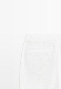 Gros plan sur un pantalon en tissu blanc avec une ceinture élastique et une poche plaquée à l'arrière sur un fond uni.