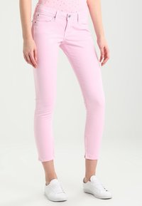 Slim-fit světle růžové džíny obuté s bílými teniskami na stojící osobě na jednobarevném světlém pozadí.