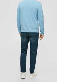 Man wearing light blue long-sleeve shirt, dark blue jeans, and white sneakers standing facing away against a plain background.