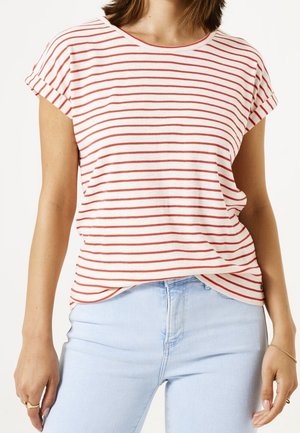 Kobieta w białym, krótkim rękawie T-shircie z cienkimi czerwonymi poziomymi paskami, włożonym w jasnoniebieskie dżinsy, z złotymi pierścionkami i bransoletką.