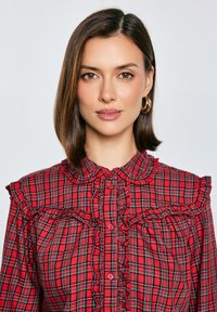 Czerwona bluzka w kratę z falbaniastym kołnierzykiem i wykończeniem z przodu, z guzikami na środku i krótkimi rękawami. Miękka bawełniana tkanina w kratkę.