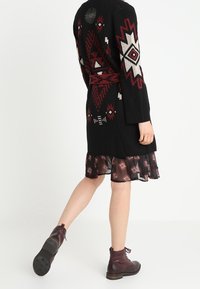 Persona che indossa un cardigan lavorato a maglia nero con motivi geometrici rossi e bianchi, una gonna trasparente a motivi e stivaletti alla caviglia marrone scuro.
