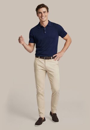 Lachende man die een navy poloshirt, beige broek, donkere riem en donkere instapschoenen draagt, met één hand op de heup staand tegen een effen achtergrond.