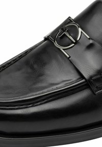 Black leather loafer with a shiny silver logo accent. Features smooth texture, stitched detailing, and a low, rounded silhouette.