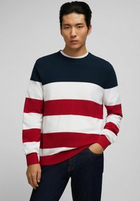 Gestreifter Pullover in Marineblau, Rot und Weiß mit Rundhalsausschnitt, strukturiertem Strickstoff und gerippten Bündchen, kombiniert mit dunklen Jeans.