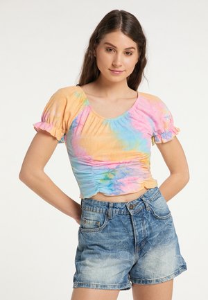 Junge Frau mit langen braunen Haaren, die ein farbenfrohes Batik-Crop-Top und hoch taillierte Jeansshorts trägt und mit den Händen in die Hüften steht.