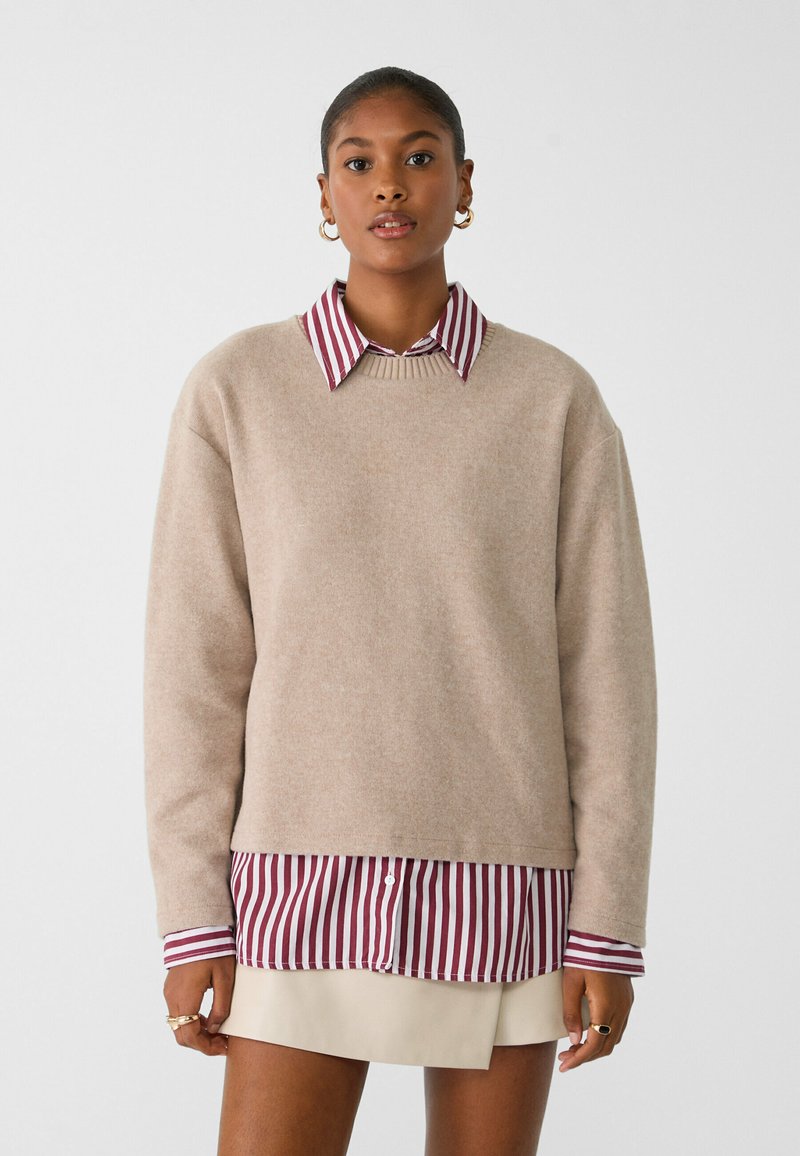 Beige trui gemaakt van zachte stof, met een ronde halslijn en verlaagde schouders, gelaagd over een bordeauxrode en witte gestreepte shirt.