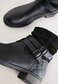Botas negras hasta el tobillo con parte superior de cuero texturizado, puño de ante, dobles hebillas y un tacón de madera bajo. Presentan detalles acolchados en el lateral.