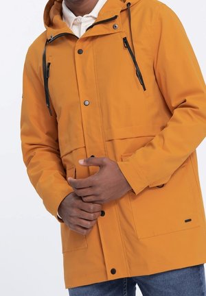 Chaqueta impermeable naranja con capucha, que incluye cierres de cremallera y a presión, cuerdas ajustables y bolsillos frontales. Textura suave.