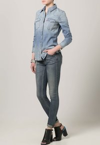 Chemise en denim bleu clair, avec deux poches poitrine et un col boutonné, associée à un jean slim bleu foncé et des sandales à talons noires.