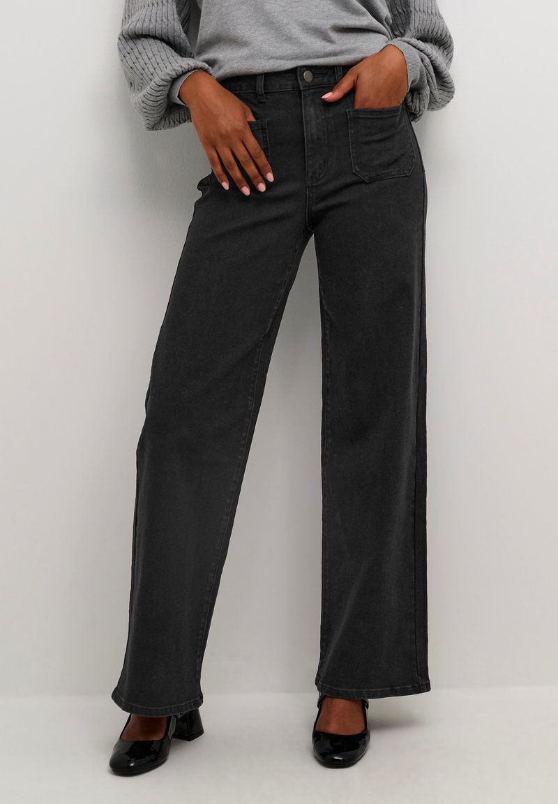 Jean noir à jambe large en denim, avec une taille haute, des poches avant et une texture lisse, associé à un pull gris et des chaussures noires.