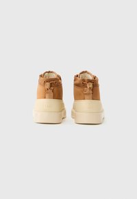 Une paire de bottes en daim beige avec des semelles en caoutchouc crème, doublure en polaire et un cordon ajustable à l'arrière, vues de l'arrière sur fond blanc.