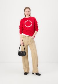 Rød hettegenser med hvit sirkulær tekst, beige vide jeans og en liten svart håndveske. Har elegante svarte sko og minimale tilbehør.